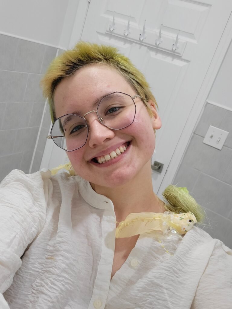 A selfie of a white person smiling, Their hair is lime green but their dark  brown roots are visible. Their hairstyle is a overgrown buzzcut. They are wearing a white pirate-esque button down, and have a fake yellow bird and green nest on their shoulder. They wear blue octagonal glasses.